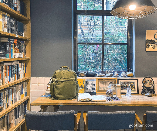 A green washed cotton canvas backpack sitting on a wooden desk in a vintage cafe next to an open book, representing the perfect analog lifestyle.