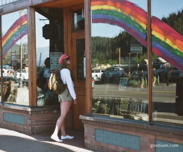 A young woman wearing a vintage olive green canvas backpack stops on a sunny street to admire a rainbow painted on a storefront window, experiencing a moment of offline freedom.