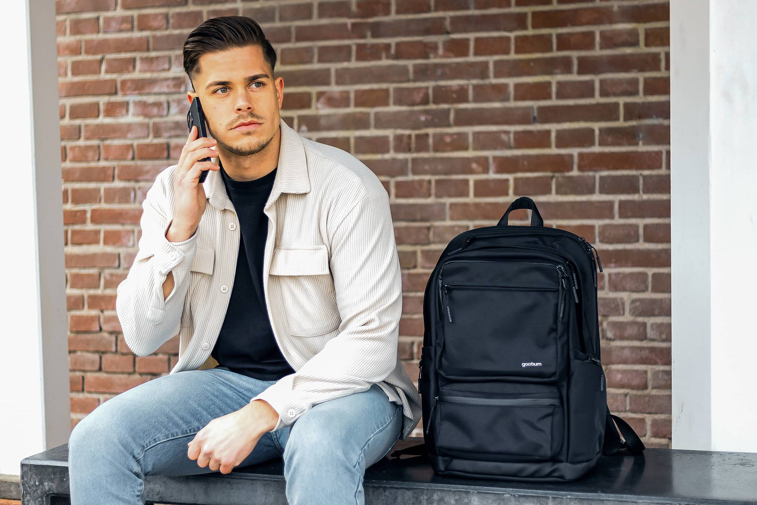 A man in a light beige shirt and blue jeans sits on a bench against a brick wall, talking on a phone next to a black laptop backpack.