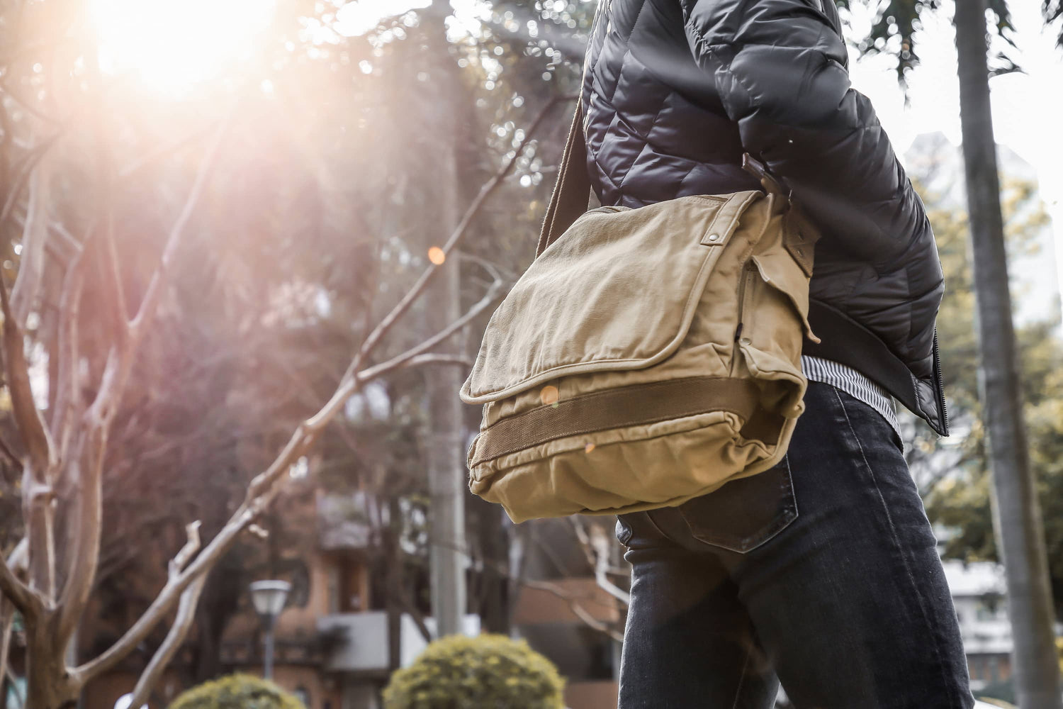 A person outdoors wearing a black puffer jacket and dark jeans, carrying a large tan canvas messenger bag over the shoulder. Sunlight is bright in the background with blurred trees and buildings.
