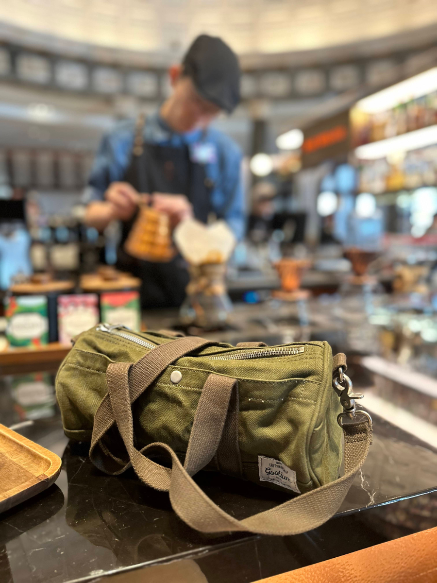 An olive green canvas crossbody bag with tan straps and a silver zipper, resting on a dark marble counter in a cafe with a blurred barista in the background.