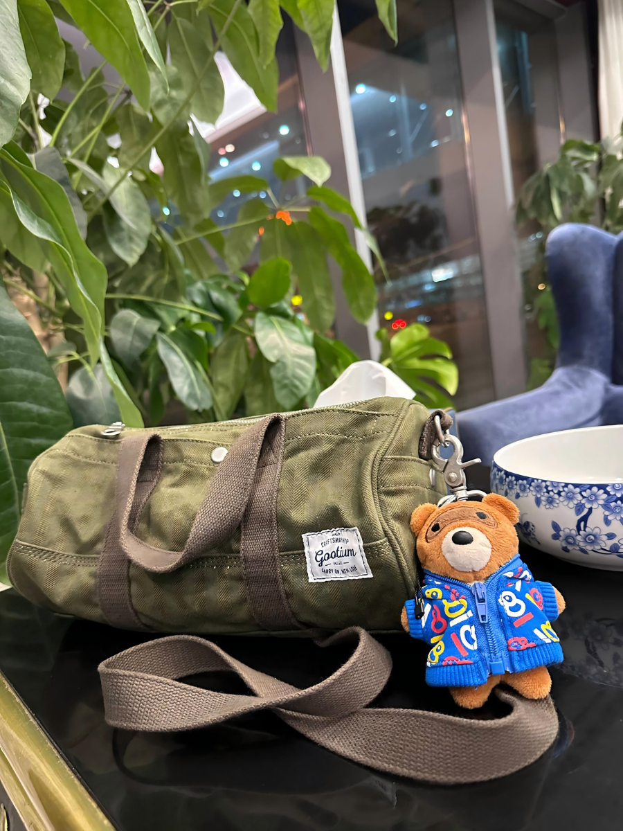 An olive green canvas mini duffel bag with brown handles and a matching shoulder strap, styled on a dark table with a small teddy bear keychain in a blue jacket. The bag has a white 'Gildan' brand label on the side.