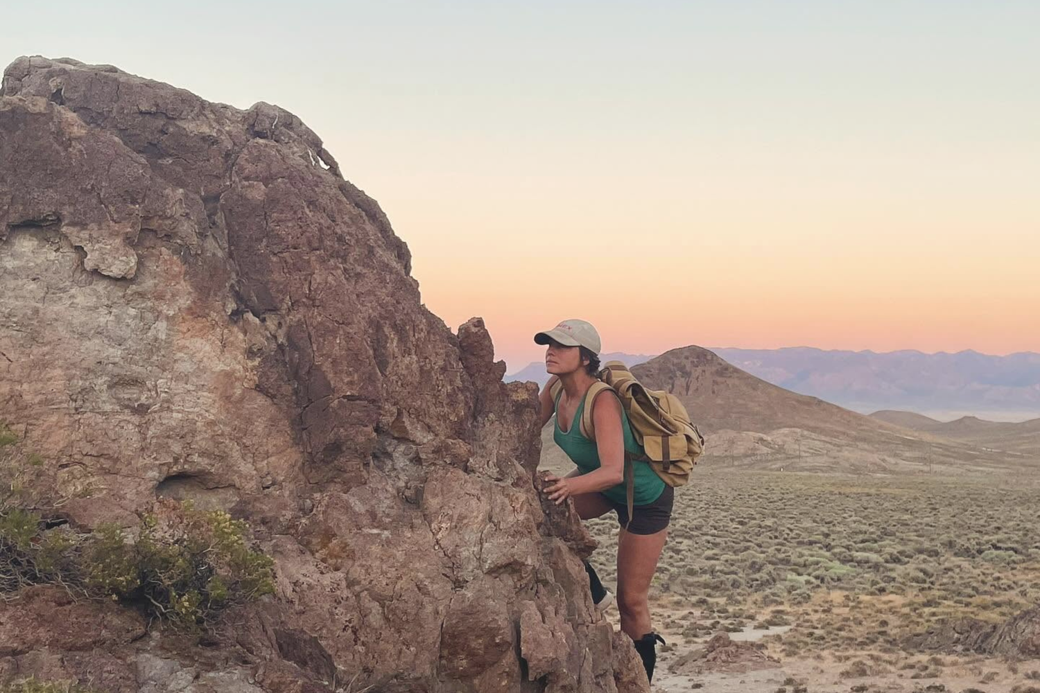 A woman hiking with a canvas backpack and cap stands against a large rock formation in a desert landscape at sunset.