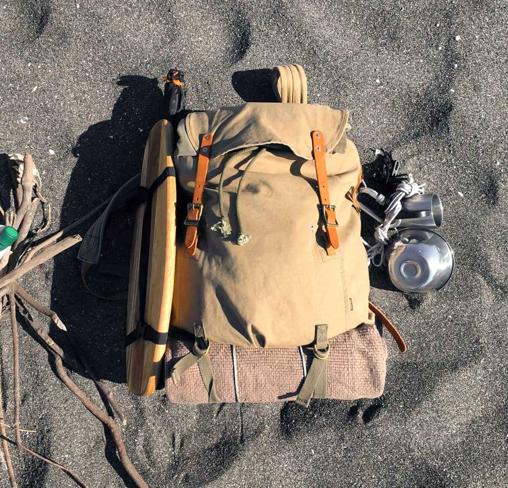 A tan canvas backpack with brown leather straps and buckles, laid flat on a dark textured surface. A rolled tan sleeping pad or blanket is underneath, with a metal canister and other gear placed to the right.