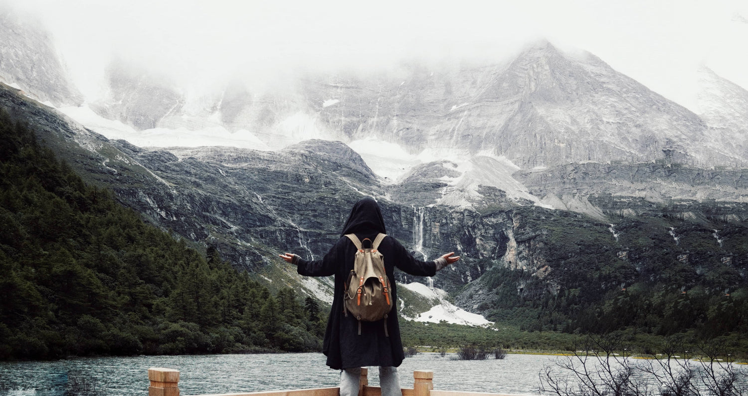 A person wearing a black hooded coat and carrying a tan backpack stands on a wooden dock with arms outstretched, facing a dramatic mountain landscape with snow, water, and mist.