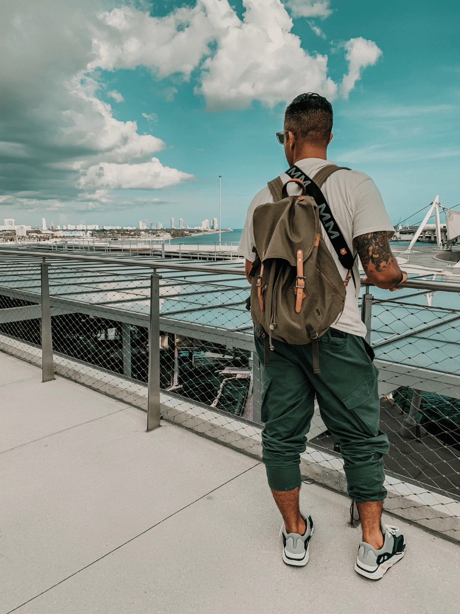 A man viewed from behind wearing a beige canvas backpack with brown leather accents, a white t-shirt, and green pants while standing on a waterfront walkway under a cloudy blue sky.