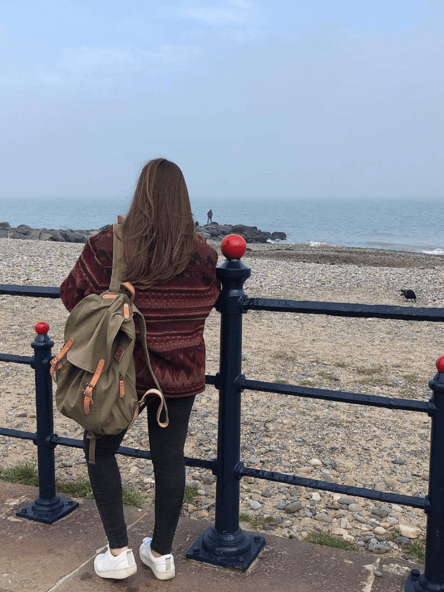A woman seen from behind wearing a burgundy striped sweater, black leggings, and white sneakers, carrying an olive green backpack with tan straps, standing on a pebble beach next to a black metal railing overlooking the sea.