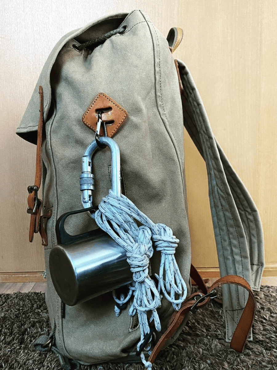 An olive green canvas backpack with brown leather accents, shown with a silver metal carabiner, a coiled blue-gray rope, and a stainless steel mug attached to its side.