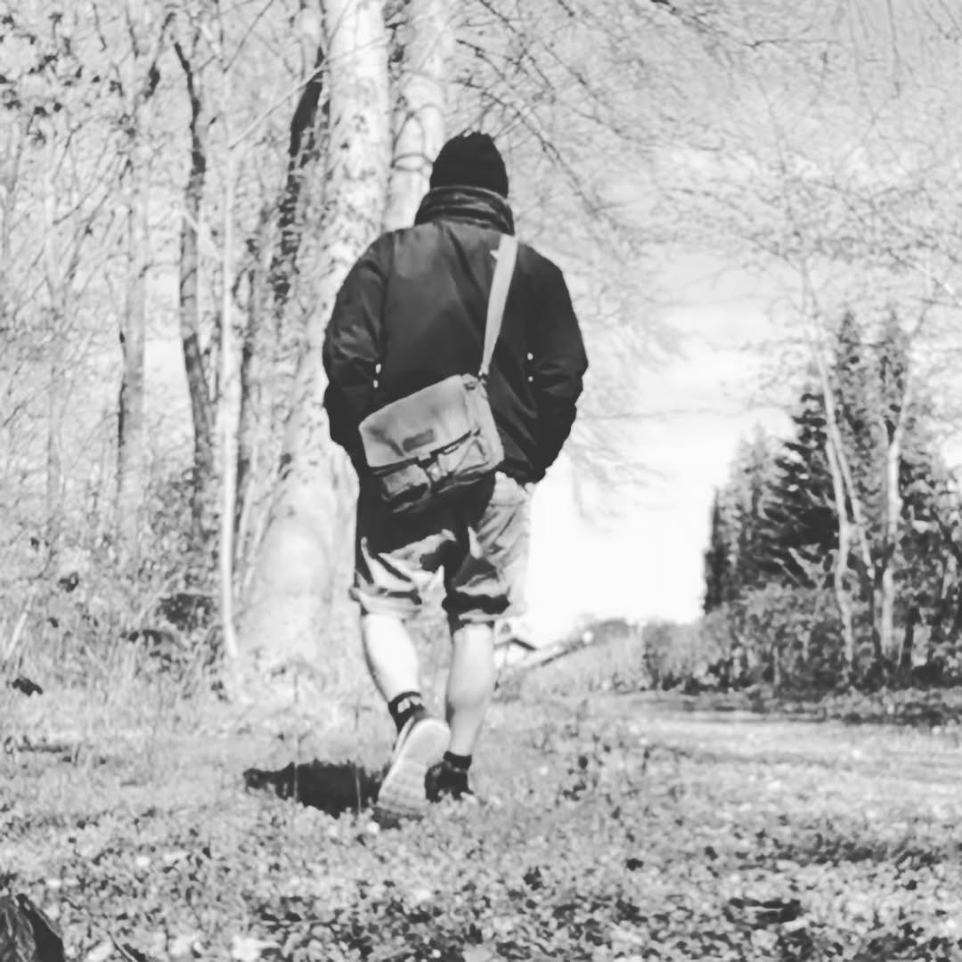A black and white photograph of a man seen from behind walking on a wooded path, wearing a dark jacket, shorts, and a crossbody messenger bag.