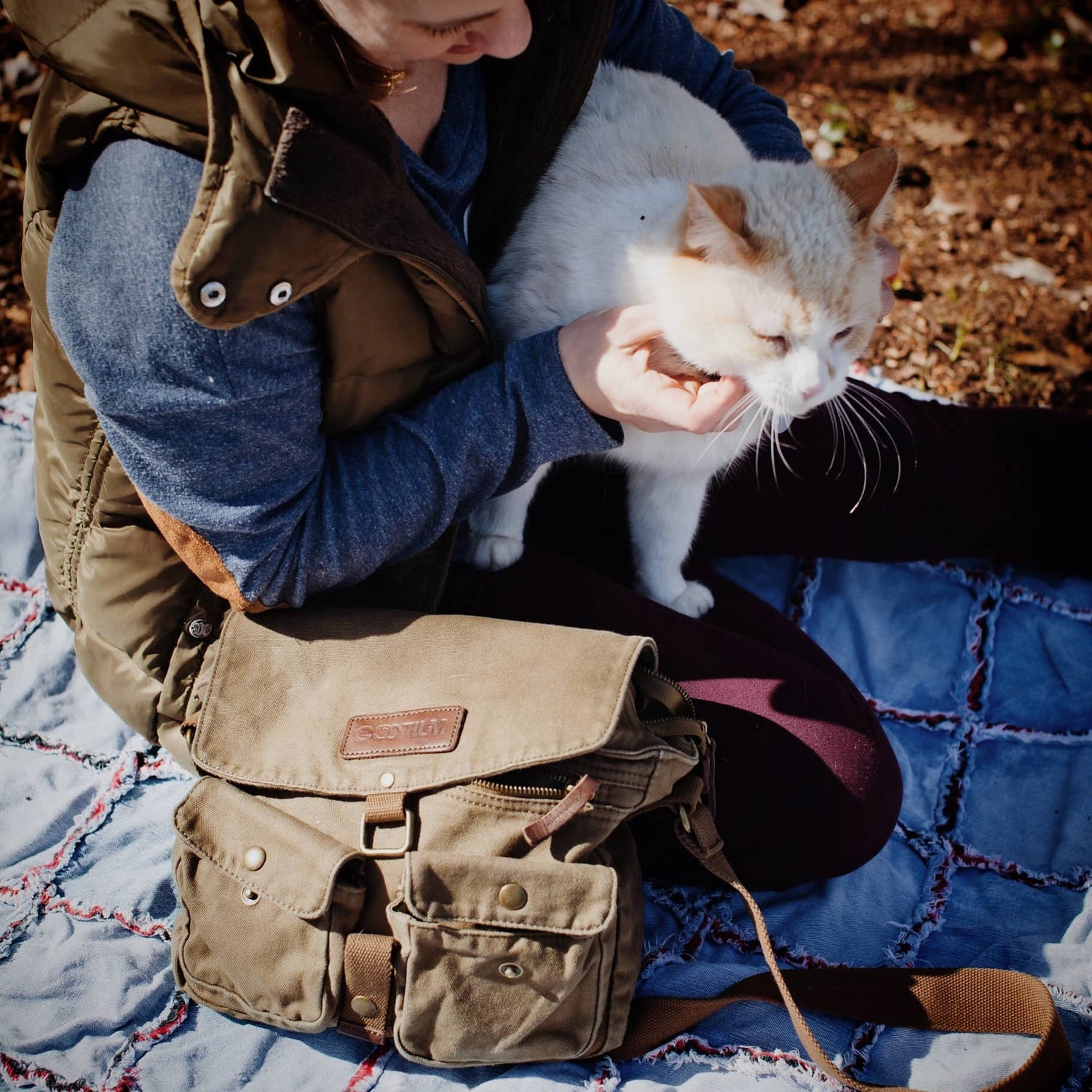 A person sitting outdoors on a blue quilted blanket while holding a white cat. A tan canvas messenger bag with brown leather trim and a small leather brand patch is resting on the lap.
