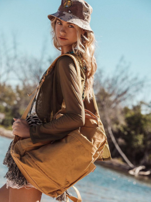 A woman outdoors by the water wearing a brown bucket hat, olive green long-sleeve top, and tan canvas backpack, photographed in warm natural light.