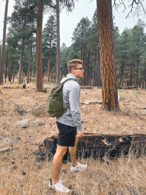 A man wearing a gray hoodie, black shorts, and white sneakers walks through a dry pine forest while wearing an olive green backpack.
