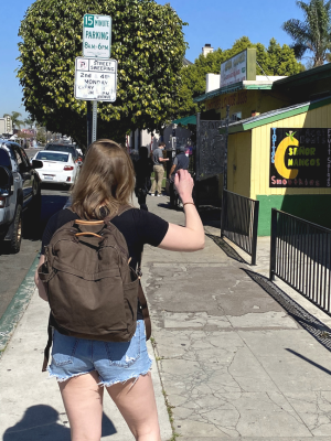 A person walking on a city sidewalk wearing a brown backpack, black t-shirt, and denim shorts, photographed from behind in an outdoor street setting.
