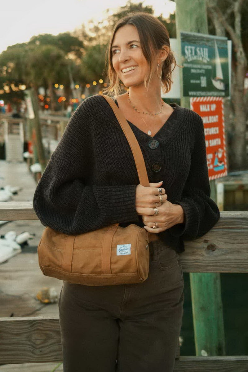A woman wearing a black knit sweater and brown pants stands on a wooden dock carrying a brown canvas shoulder bag with a small white brand patch. The background shows a blurred outdoor harbor scene at sunset.