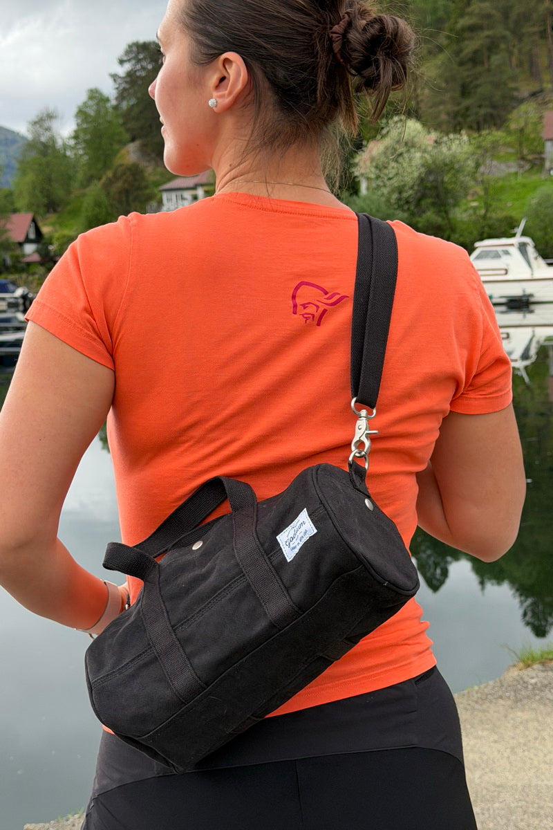 A woman wearing an orange t-shirt and black pants is shown from behind, carrying a black canvas crossbody duffel bag with a silver clasp and a small white brand label. The background shows a scenic outdoor view of water, trees, and boats.