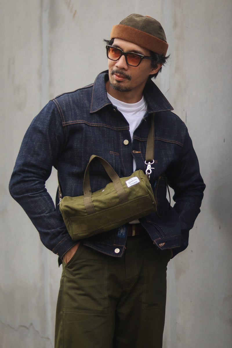 A man wearing a dark blue denim jacket, olive green cargo pants, a brown knit beanie, and orange-tinted sunglasses, carrying an olive green canvas crossbody bag across his chest against a textured gray wall.