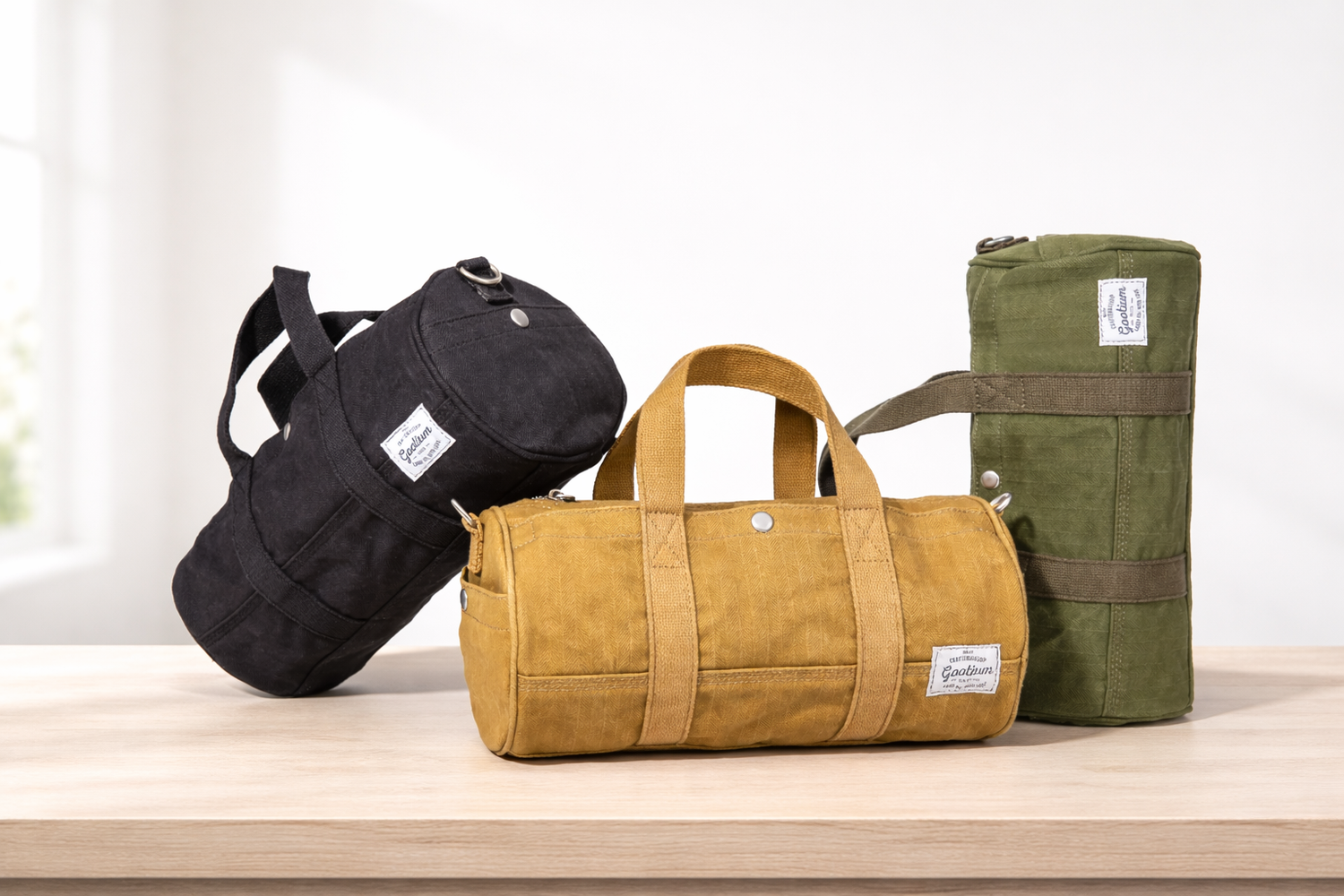 Three canvas barrel-style bags displayed on a light wood surface against a bright white background: a black bag leaning on the left, a mustard yellow bag centered in front, and an olive green bag standing on the right. Each bag has a small rectangular brand patch on the front.