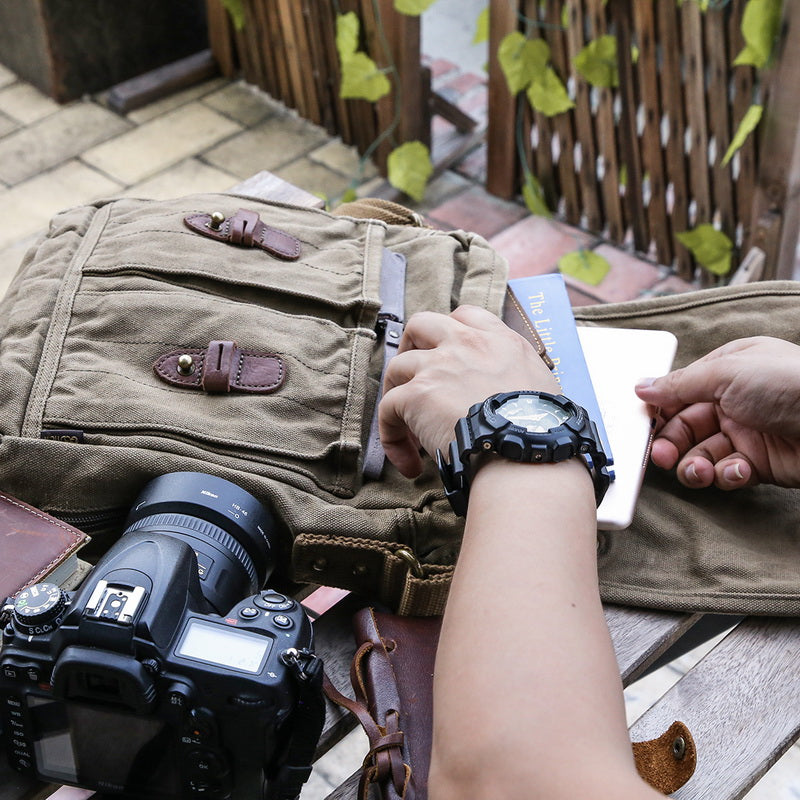 A close-up lifestyle shot of a person's hands placing a passport into the pocket of a khaki canvas satchel with brown leather straps. A black camera is visible in the foreground, and the setting is outdoors on a wooden table.