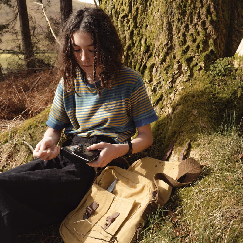 A person sitting outdoors against a mossy tree, holding a smartphone and wearing a blue-and-yellow striped t-shirt and black pants. A tan canvas satchel lies on the grass beside them in a wooded setting.