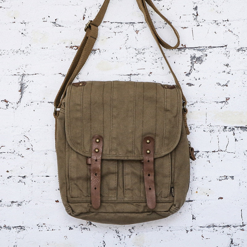 An olive green canvas messenger bag with brown leather straps and brass hardware, hanging against a distressed white wooden wall.