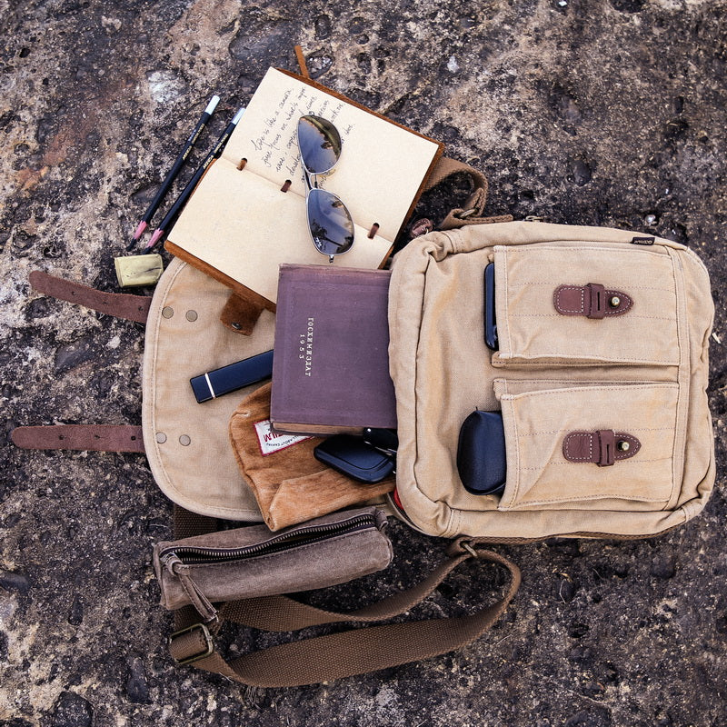 A beige canvas crossbody bag laid open on a rough stone surface, styled with an open notebook, sunglasses, a purple book, a small pouch, and other travel accessories. No person or animal is present, but this is not a clean isolated studio shot; it is a styled flat-lay product presentation, so Product/Packaging fits better than Lifestyle.