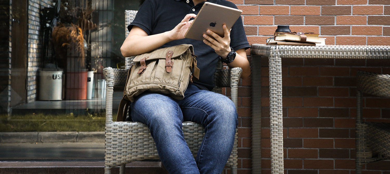 A person sitting in a wicker chair by a brick wall, using a tablet while wearing a tan canvas messenger bag across their lap. They are dressed in a black t-shirt and blue jeans.