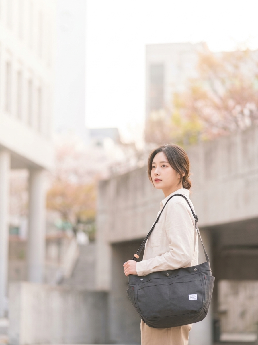 A woman wearing a beige cardigan and beige pants carries a black canvas shoulder bag with a white label, standing outdoors in front of a concrete structure with soft-focus cherry blossoms.