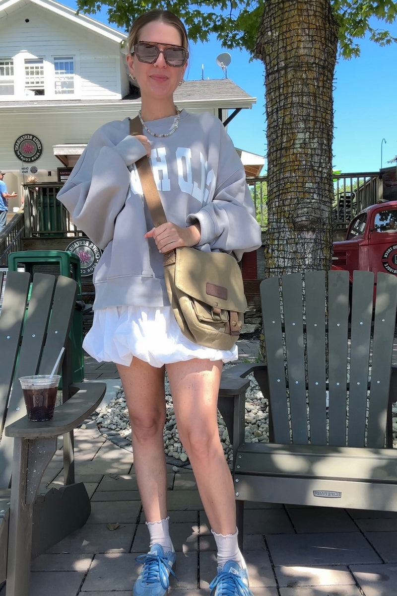 A woman stands outdoors in a cafe or patio setting wearing a green canvas messenger bag with 'GOOTIUM' printed on the front, a white ruffled mini skirt, blue sneakers, and carrying a tan messenger bag.