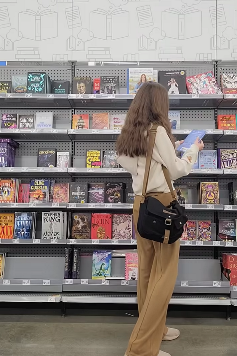 A woman with long brown hair stands in a bookstore aisle, viewed from behind as she looks at books on a shelf. She is wearing a cream long-sleeve top, tan wide-leg pants, and a black canvas shoulder bag.