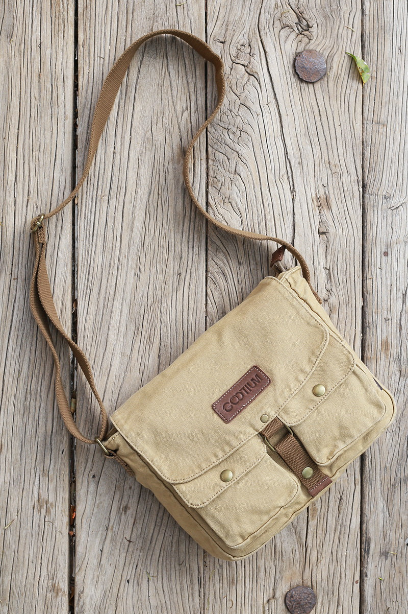 A beige canvas crossbody bag with brown leather accents and a long shoulder strap, laid flat on a rustic weathered wood plank surface.
