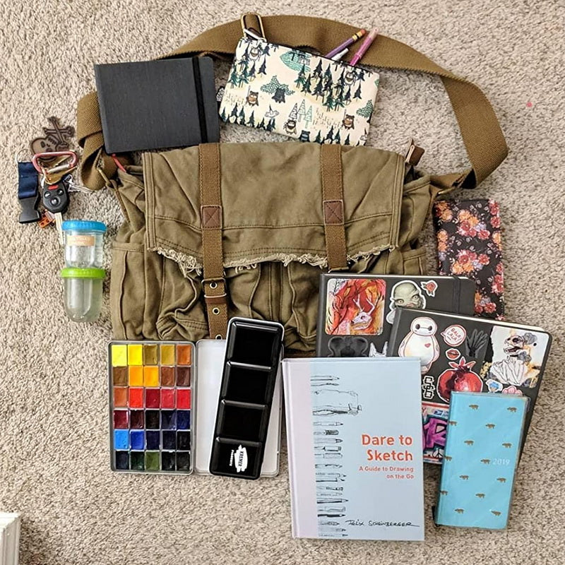 An overhead flat-lay of a khaki canvas messenger bag with its strap, surrounded by sketching and travel accessories on a beige carpet background. The bag is styled with a patterned pouch, notebook, water bottle, paint palette, phone case, stickers, and sketchbooks.