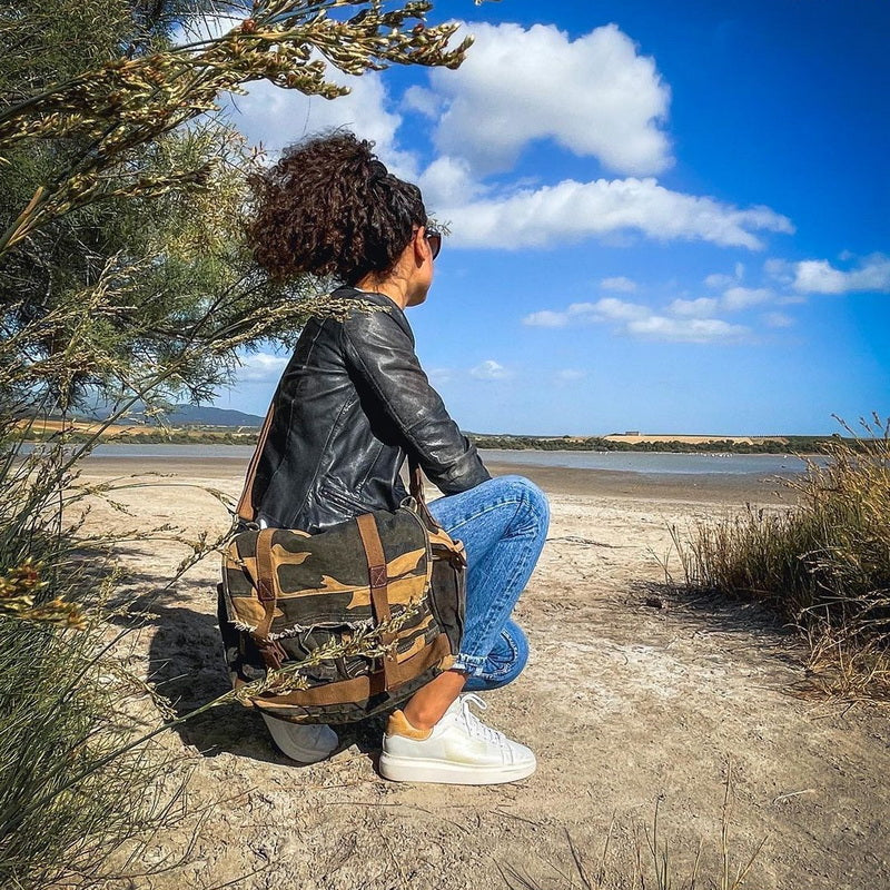 A person crouches outdoors by a sandy shoreline under a bright blue sky, wearing a black leather jacket, blue jeans, and white sneakers, while carrying a large camouflage shoulder bag.