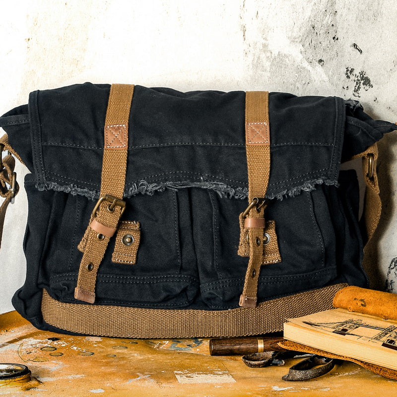 A black canvas messenger bag with tan webbing straps and leather buckles, resting on a rustic wooden table against a textured plaster wall.