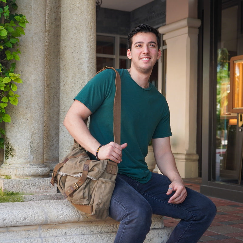 A smiling young man sits on stone steps in front of a building, wearing a green t-shirt, dark jeans, and carrying a tan canvas messenger bag with brown leather straps.