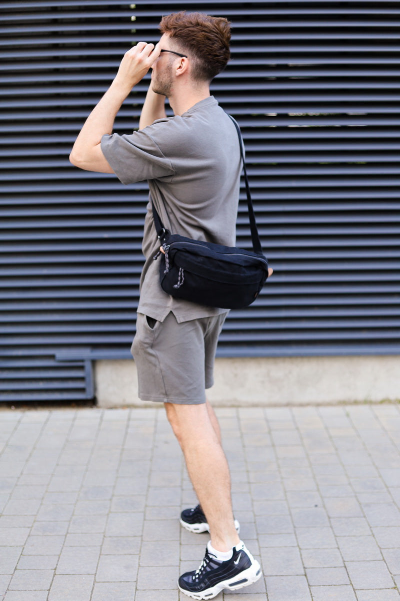 A man wearing a gray t-shirt and shorts, carrying a black crossbody sling bag, standing in front of a dark gray slatted wall.
