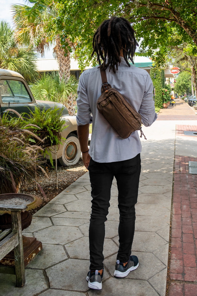 A man viewed from behind wearing a brown crossbody sling bag, a light gray button-up shirt, black pants, and sneakers, walking on a stone sidewalk outdoors.