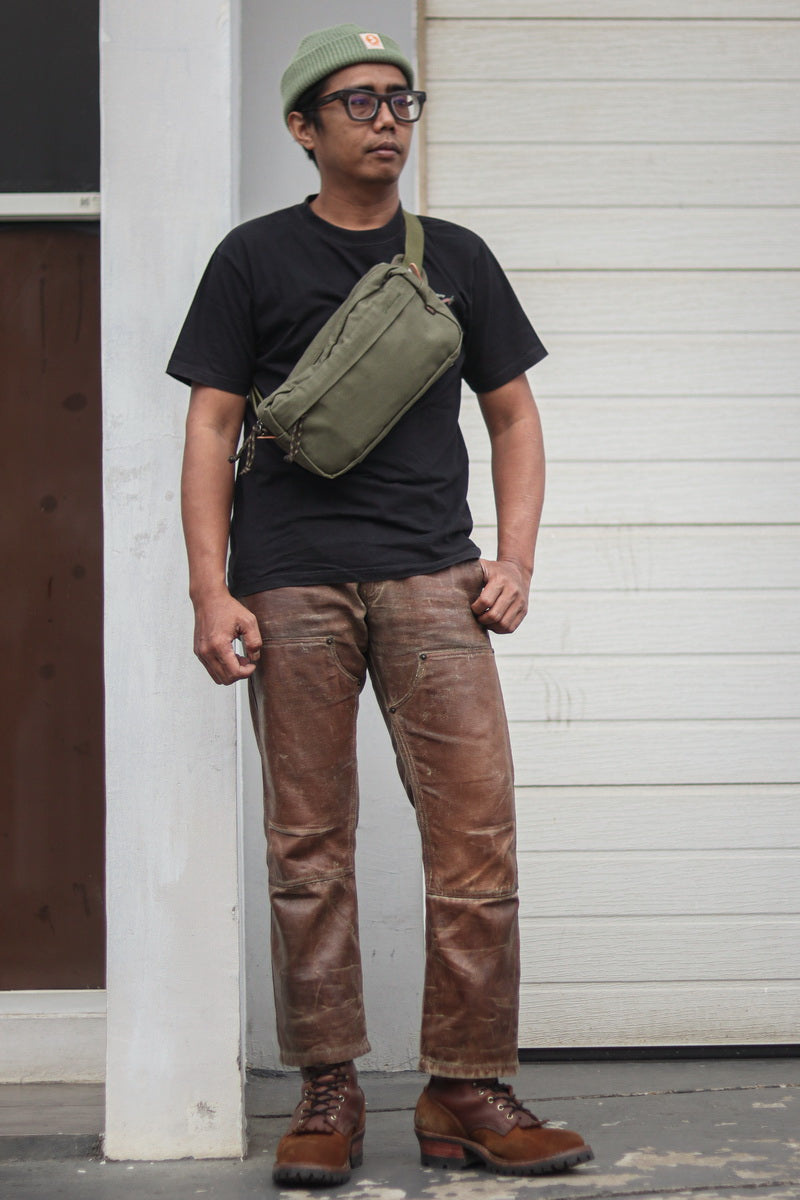 A man wearing a black t-shirt, brown distressed jeans, brown boots, a green beanie, and glasses, carrying an olive green crossbody sling bag against a white wooden door background.