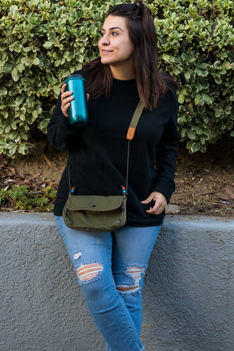 A woman wearing a black crewneck sweatshirt, ripped blue jeans, and a small olive green crossbody bag, holding a teal tumbler against a background of green bushes and a gray wall.