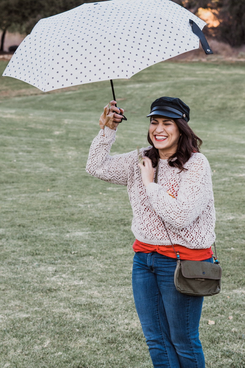 A woman standing on a grassy lawn holds up a white umbrella with black polka dots. She is wearing a cream-colored crochet knit sweater, a bright orange shirt underneath, blue jeans, a black cap, and carrying a small olive green crossbody bag.