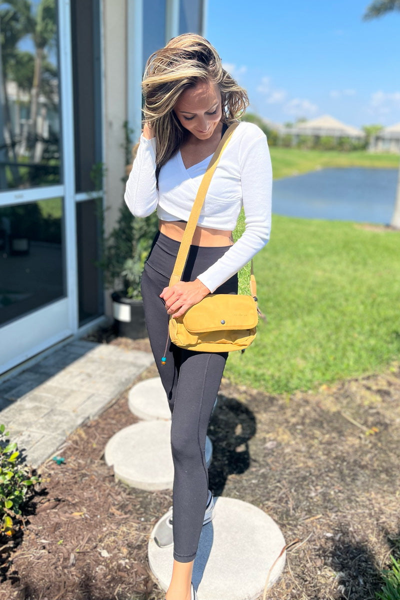 A woman standing outdoors on stone pavers near a house, wearing a white long-sleeve crop top, dark gray leggings, and carrying a mustard yellow crossbody bag.