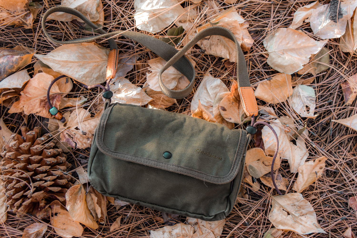 An olive green canvas crossbody bag with a flap closure and tan leather accents, laid on a bed of dry autumn leaves and pine needles outdoors.