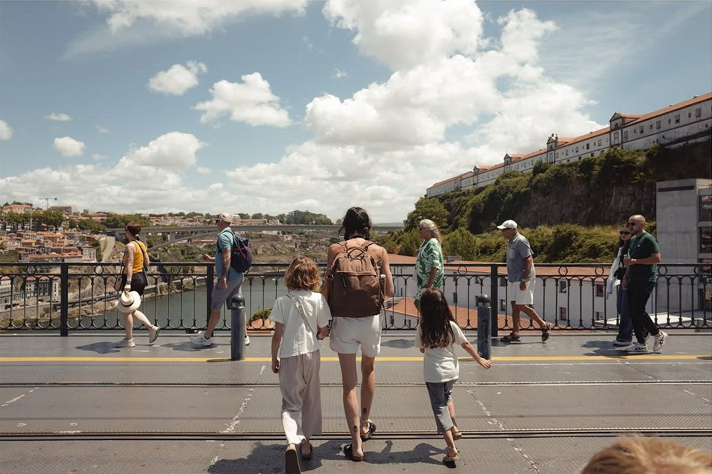 A family of four, including a woman wearing a canvas backpack and two children, walks away from the camera on a city bridge, with other pedestrians and a scenic river valley and historic buildings in the background under a partly cloudy sky.