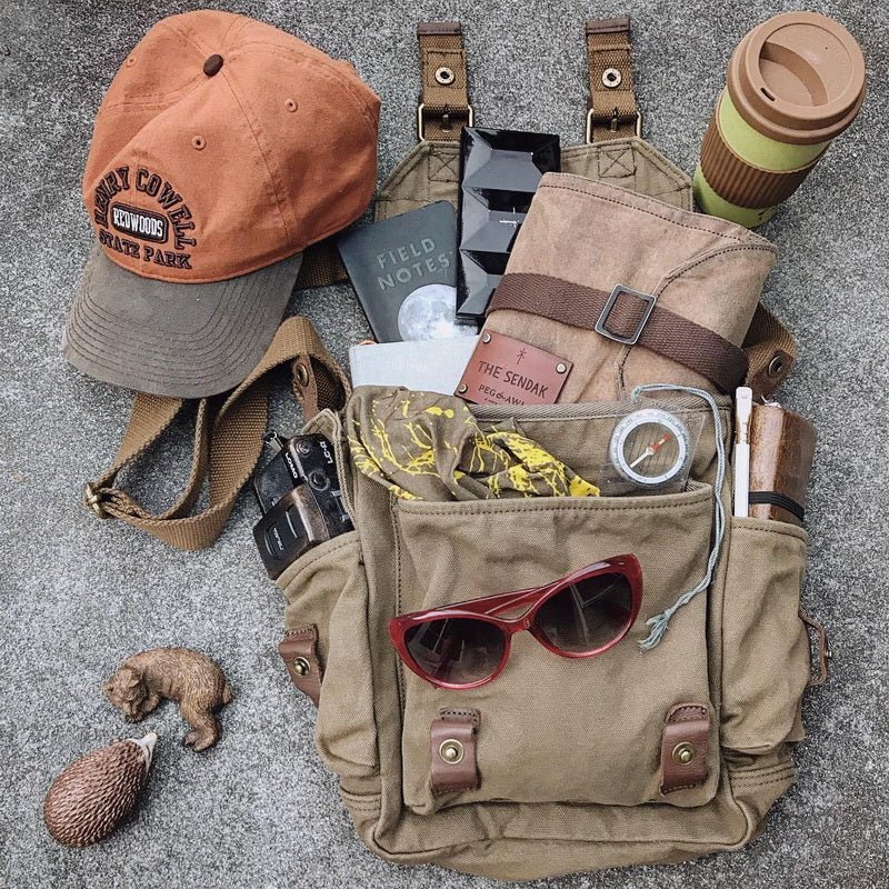 A styled flat-lay of a tan canvas messenger bag filled with everyday carry items, including an orange baseball cap, a brown travel mug, sunglasses, tape, keys, and a small hedgehog figurine, arranged on a gray textured surface.