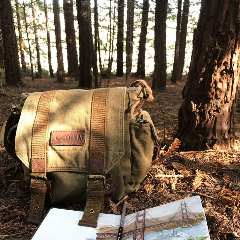 A tan canvas messenger bag with brown leather straps and a 'COOTIUM' brand patch, set on a forest floor surrounded by pine trees, with an open notebook in the foreground.