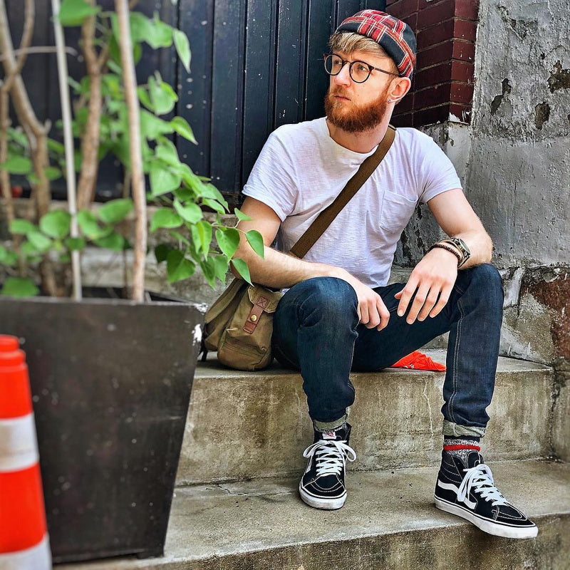 A man with a beard and glasses sits on concrete steps wearing a white t-shirt, dark blue jeans, black and white high-top sneakers, a red plaid beanie, and a brown canvas messenger bag slung over his shoulder.