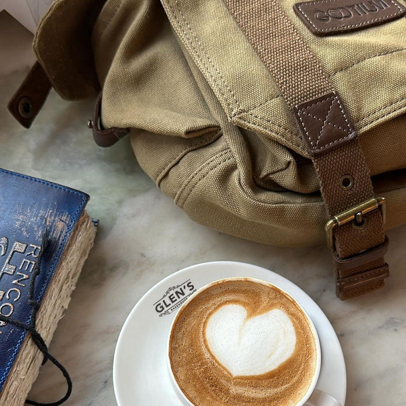 A tan canvas messenger bag with brown leather straps and brass hardware, styled on a light marble surface next to a cup of latte with heart-shaped foam art and a blue notebook.