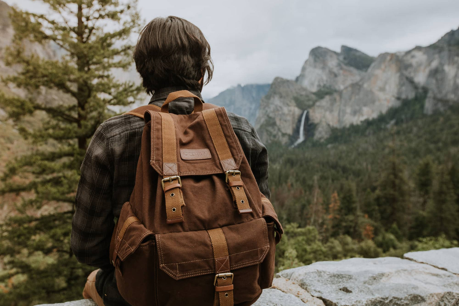 A person seen from behind wearing a brown canvas backpack with tan leather straps, sitting outdoors in a mountain landscape with trees and a waterfall in the background.