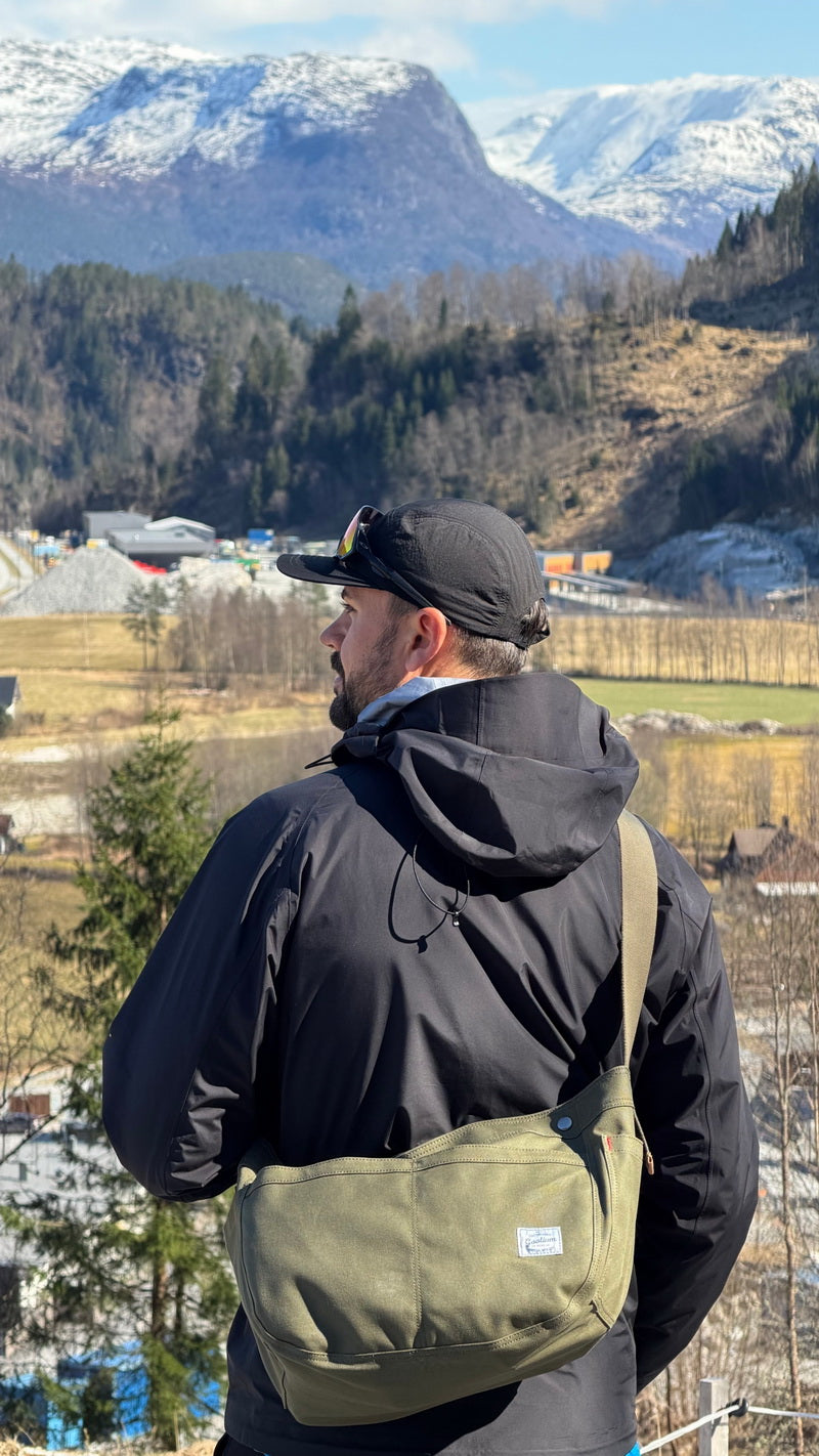 A man wearing a black jacket and cap, viewed from behind, wearing an olive green crossbody messenger bag with a small white label. He is standing outdoors in front of a scenic mountain landscape with snow patches and trees.