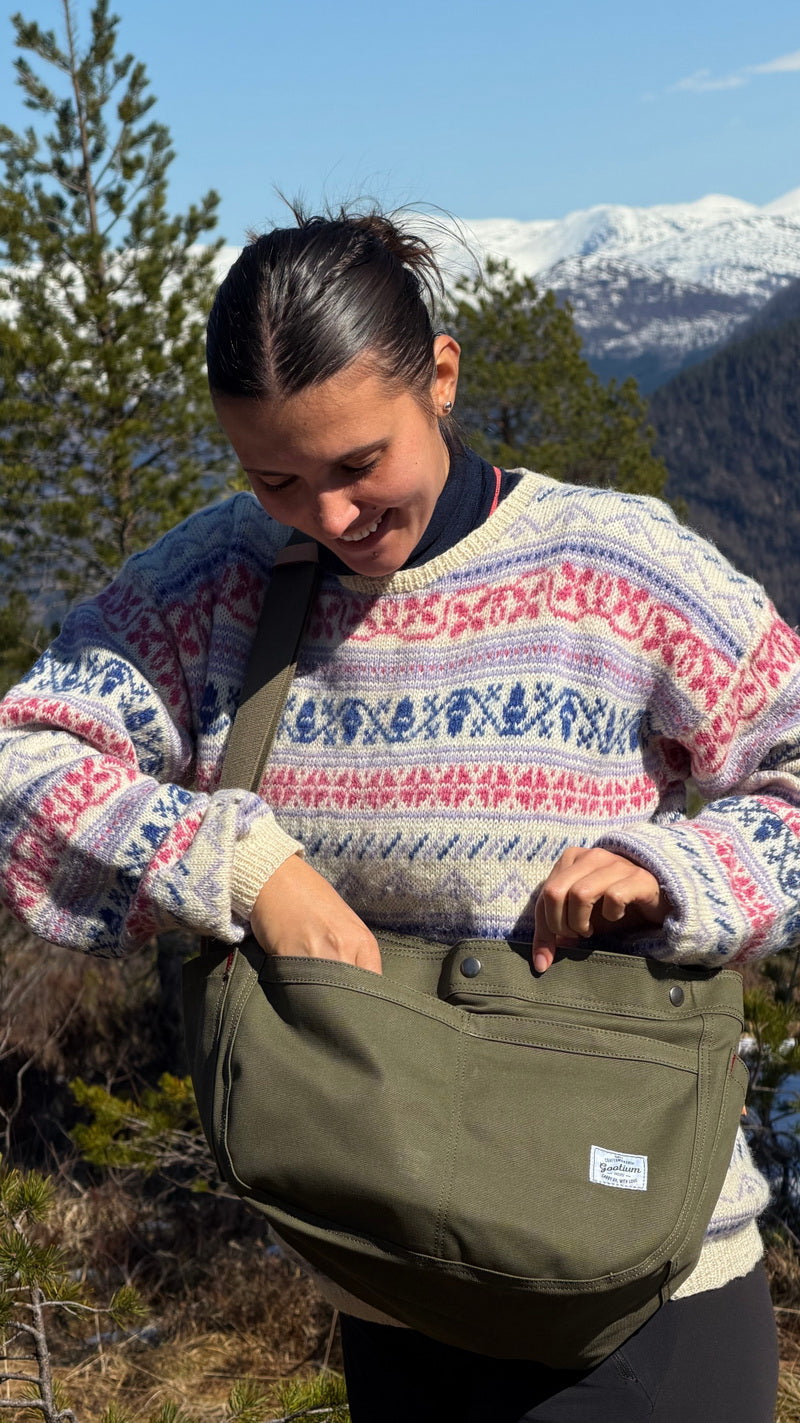 A woman wearing a colorful patterned knit sweater stands outdoors in a mountain landscape, holding and looking into an olive green canvas shoulder bag.