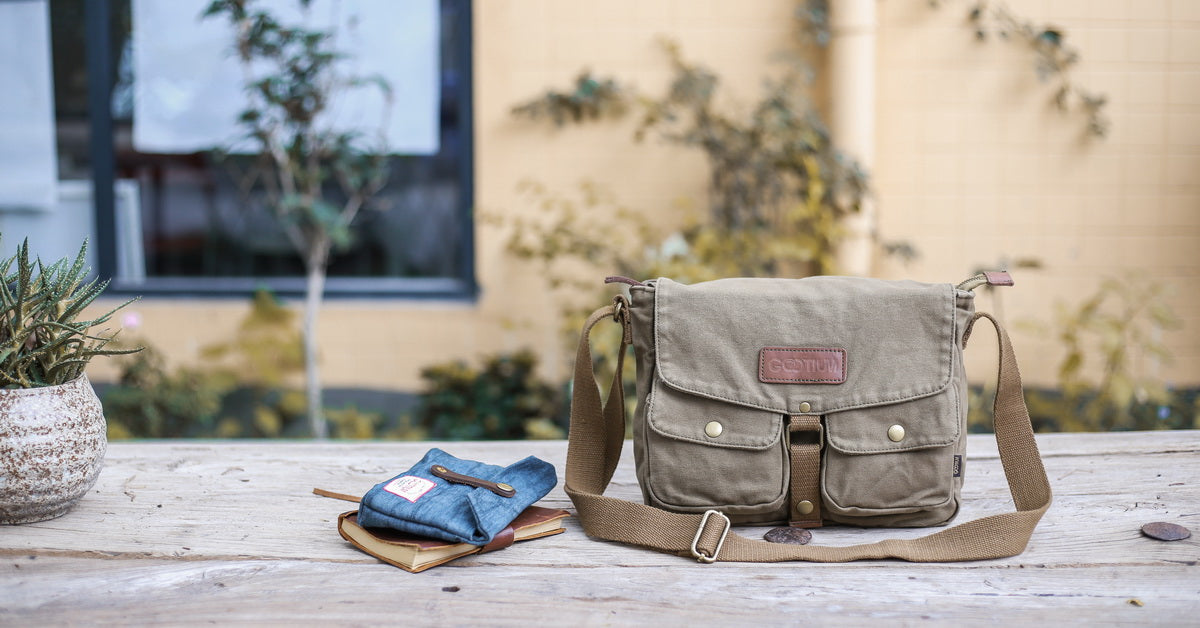 An olive green canvas messenger bag with brown leather trim and a brown leather brand patch, resting on a rustic wooden table next to a blue denim pocket square. The background shows a softly blurred beige building wall with green foliage.